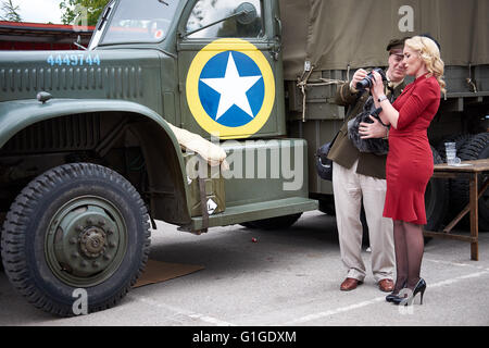 Man dressed in vintage soldier US military uniform shows camera to blonde woman dressed in red dress with black high heels Stock Photo