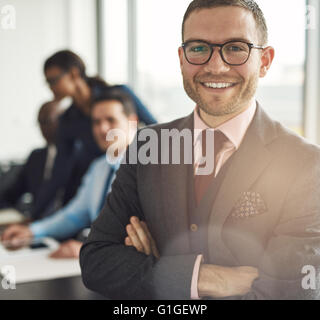Smiling confident businessman wearing glasses standing with folded arms looking at the camera, close up view with colleagues wor Stock Photo