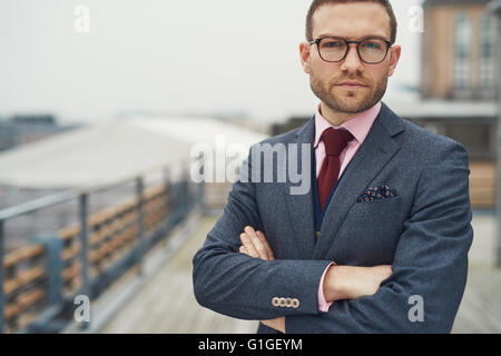 Stylish serious handsome bearded young businessman in eyeglasses, red necktie and folded arms outside on balcony near railing Stock Photo