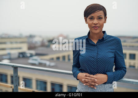 Single calm business woman with folded hands leaning against railing outside with overcast urban skyline and copy space in backg Stock Photo