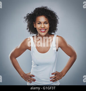 Front centered view on happy young Black female with enthusiastic expression leaning forward with hands on hips over gray backgr Stock Photo