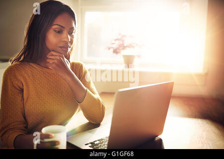 Serious beautiful woman with hand on chin sitting near bright window while looking at open laptop computer on table and holding Stock Photo