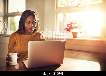 Adult female holding cup and laughing while looking at her laptop computer while sitting near bright sunny window in kitchen Stock Photo