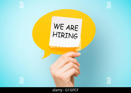 Adhesive note on yellow speech bubble with "we are hiring" words on blue background. Stock Photo