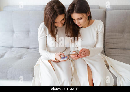 Two concentrated pretty young sisters twins sitting on couch and using their mobile phones Stock Photo