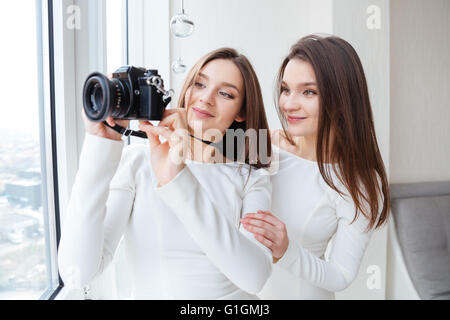 Two happy cute sisters twins taking photos using camera near the window Stock Photo