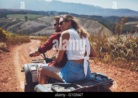 Rear view shot of young couple riding on a quad bike in countryside and looking away smiling. Woman sitting behind her boyfriend Stock Photo