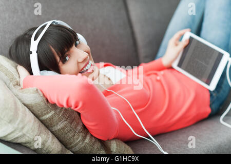 Smiling young woman relaxing at home on the couch, she is wearing headphones, using a digital tablet and watching a video online