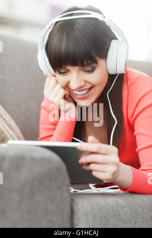 Smiling young woman relaxing at home on the couch, she is wearing headphones, using a digital tablet and watching a movie online