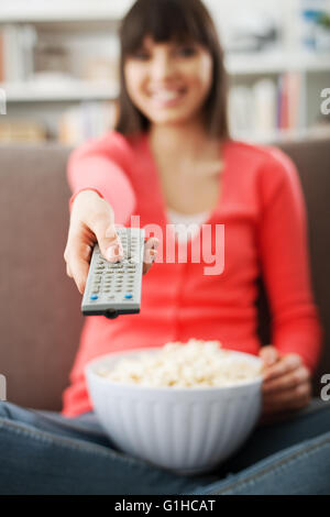 Young smiling woman at home sitting on the couch and watching tv, she is holding a remote control and eating popcorn