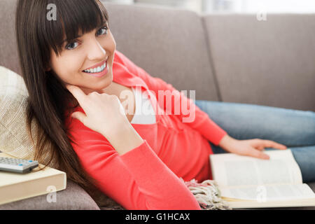 Beautiful young woman relaxing at home on the sofa and reading a book, she is smiling at camera Stock Photo