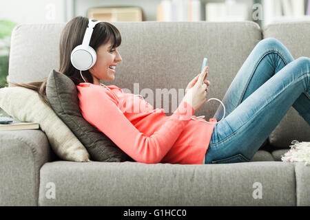 Attractive young woman on the sofa at home, she is playing music with her smarphone and wearing headphones, leisure and entertai Stock Photo