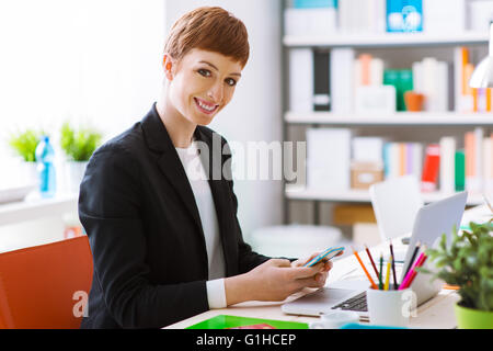 Successful confident businesswoman in her office using a smart phone, she is texting and using a mobile app Stock Photo