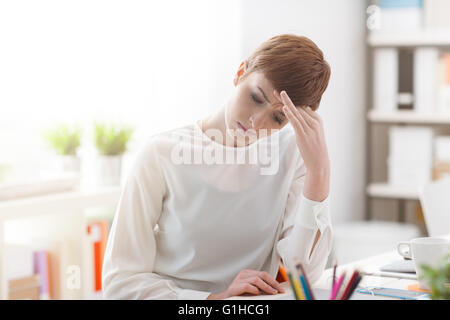 Tired businesswoman sitting at office desk and having an headache, she is touching her forehead, stress and disease concept Stock Photo