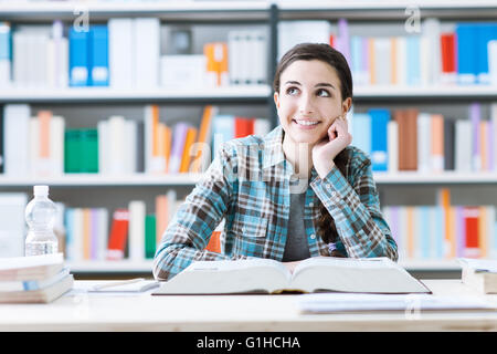Smiling student girl in the library studying and day dreaming, she is thinking with hand on chin and looking up, education and Stock Photo