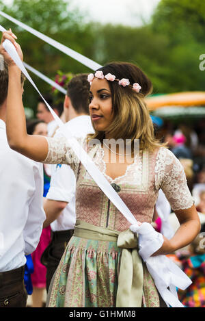 Portrait of a dark-skinned young woman in a dress dancing a traditional Bavarian folk dance around a maypole holding a ribbon Stock Photo