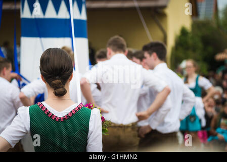 Woman in local dress watching young men in Lederhose standing around a maypole while dancing a traditional Bavarian folk dance Stock Photo