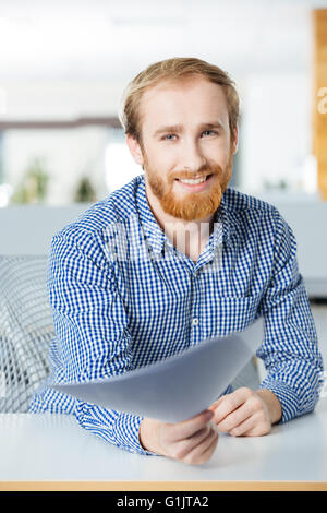 Cheerful bearded young businessman sitting in office and giving papers to you Stock Photo