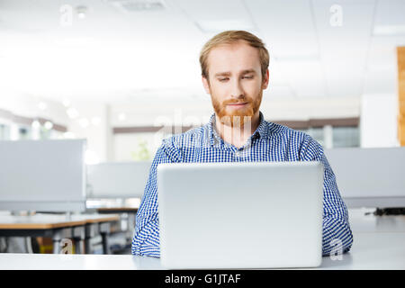 Attractive focused bearded young businessman sitting and using laptop on workplace in office Stock Photo