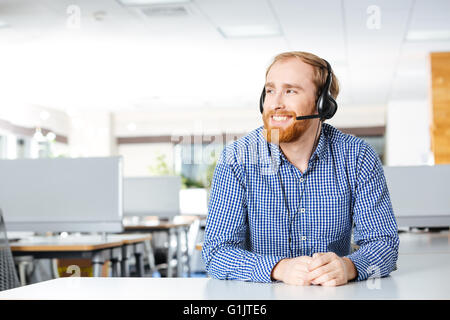 Cheerful bearded young man sitting and using headset in office Stock Photo