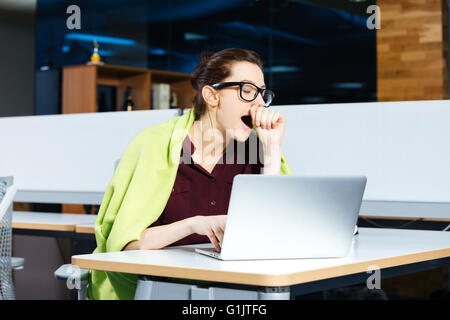 Pretty overworked young businesswoman in glasses yawning and using laptop on workplace Stock Photo
