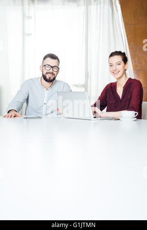 Portrait of two smiling attractive young businesspeople sitting at the table and using laptop in meeting room Stock Photo
