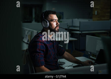 Concentrated handsome young man in headphones sitting and working at night in dark office Stock Photo