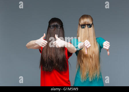 Two comical young women with faces covered by long hair showing thumbs up and thumbs down over grey background Stock Photo