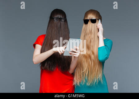 Two amusing young women with faces covered by long hair using tablet and cell phone over grey background Stock Photo