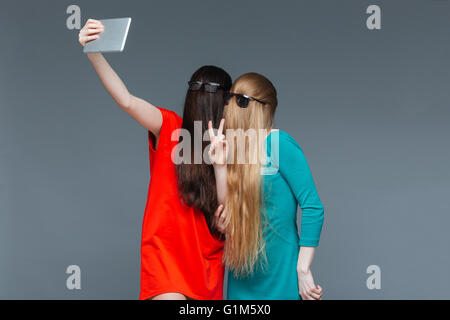 Two comical young women with faces covered by long hair taking selfie with tablet over grey background Stock Photo
