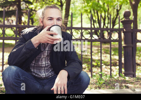 Bearded Asian man sitting on the sidewalk in park and drinking coffee from paper cup Stock Photo
