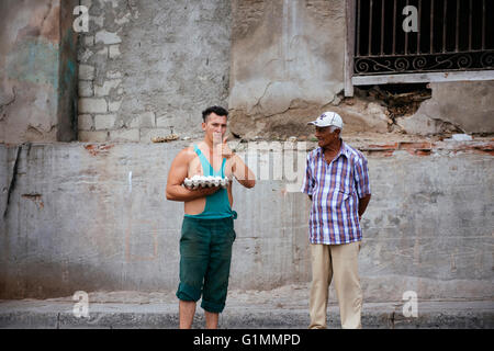 A young Cuban man is holding a tray of fresh eggs and giving a thumbs up while an elderly Cuban man watches him in Santiago de Cuba, Cuba. Stock Photo