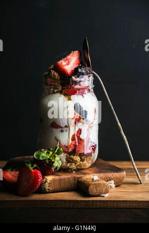 Yogurt oat granola with strawberries, mulberries, honey and mint leaves in tall glass jar on black backdrop Stock Photo