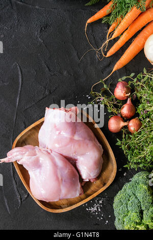 Raw uncooked rabbit legs in wooden plate ready to cook with vegetables, served over black textured background. Top view. Copy sp Stock Photo