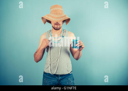A happy young man wearing a beach hat and pearl necklaces is drinking from a blue cup Stock Photo