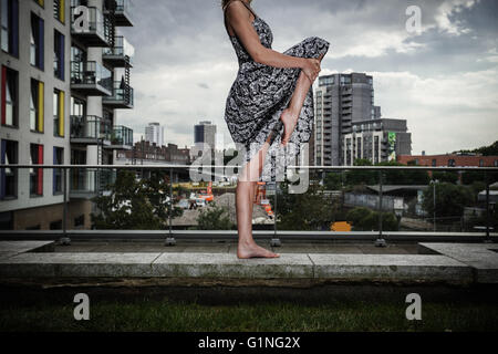 A young woman wearing a dress is raising her leg as she is standing on a roof top Stock Photo