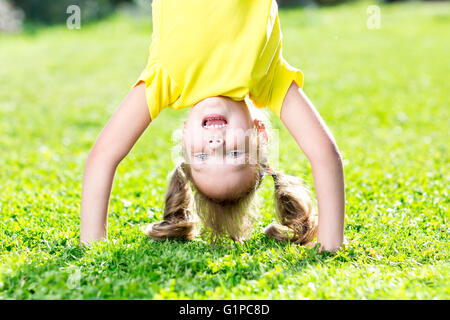 Happy little child girl have a fun standing on her head among grass Stock Photo