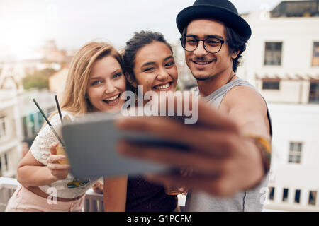 Shot of young man with female friends taking selfie on mobile phone. Young people partying on rooftop having fun. Stock Photo