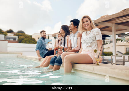 Portrait of happy young woman sitting by the pool with her friends partying. Young people relaxing by the swimming pool with the Stock Photo