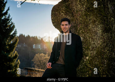 Handsome young man leaning against tree, looking at camera, in a sunny day wearing a black coat Stock Photo