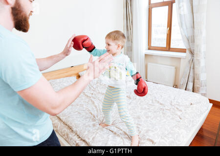 Smiling little boy in red boxing gloves standing on bed and playing with his dad at home Stock Photo