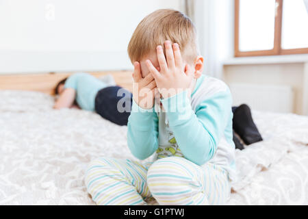 Sad little boy sitting and crying on bed while his father is sleeping Stock Photo