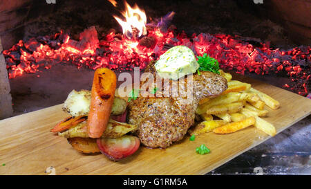 Ox, beef or cow minute steak portion, featuring seasoned butter, french fries and grilled vegetables, in front of a stone oven f Stock Photo