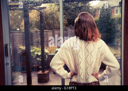 A young woman is standing by the french doors in her house and is looking out at the rain Stock Photo