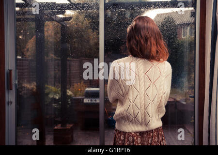 A young woman is standing by the french doors in her house and is looking out at the rain Stock Photo