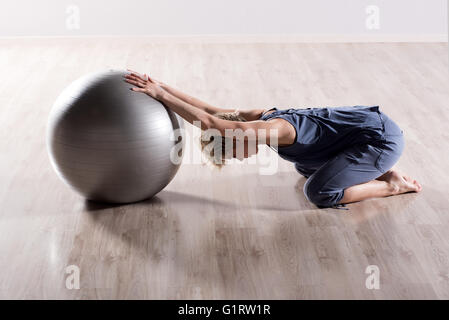 Single flexible woman stretching her shoulders with outstretched arms on top of silver fitness ball on hardwood studio floor Stock Photo