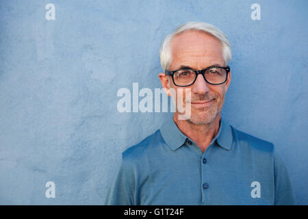 Close up portrait of handsome senior man with glasses looking at camera, he is standing against a blue wall. Stock Photo