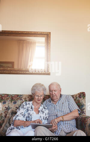 Vertical indoor shot of retired couple at home using digital tablet. Senior caucasian man and woman sitting together on sofa at Stock Photo