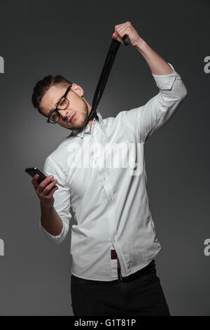 Sad hopeless young businessman using mobile phone and choking himself with his own tie over grey background Stock Photo