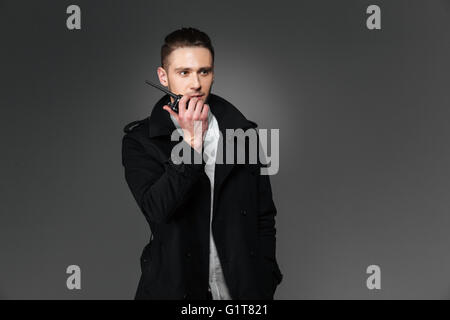 Serious young man in black coat talking in walkie talkie over grey background Stock Photo